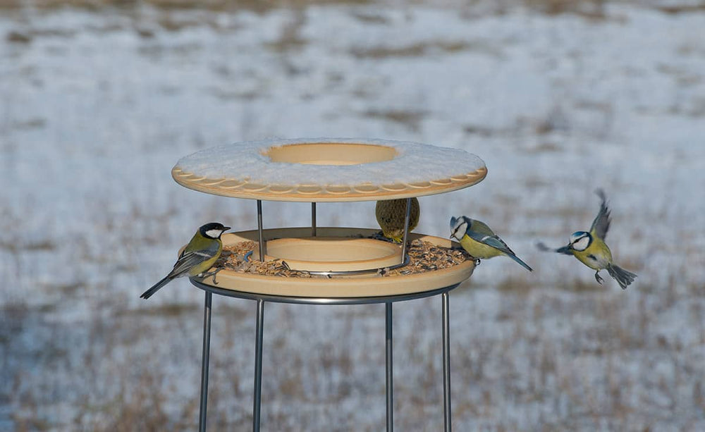 Vogelfutterhaus CeraNatur auf Stahlgestell, rund, schneebedeckt, umringt von mehreren Vögeln in einer winterlichen Umgebung.