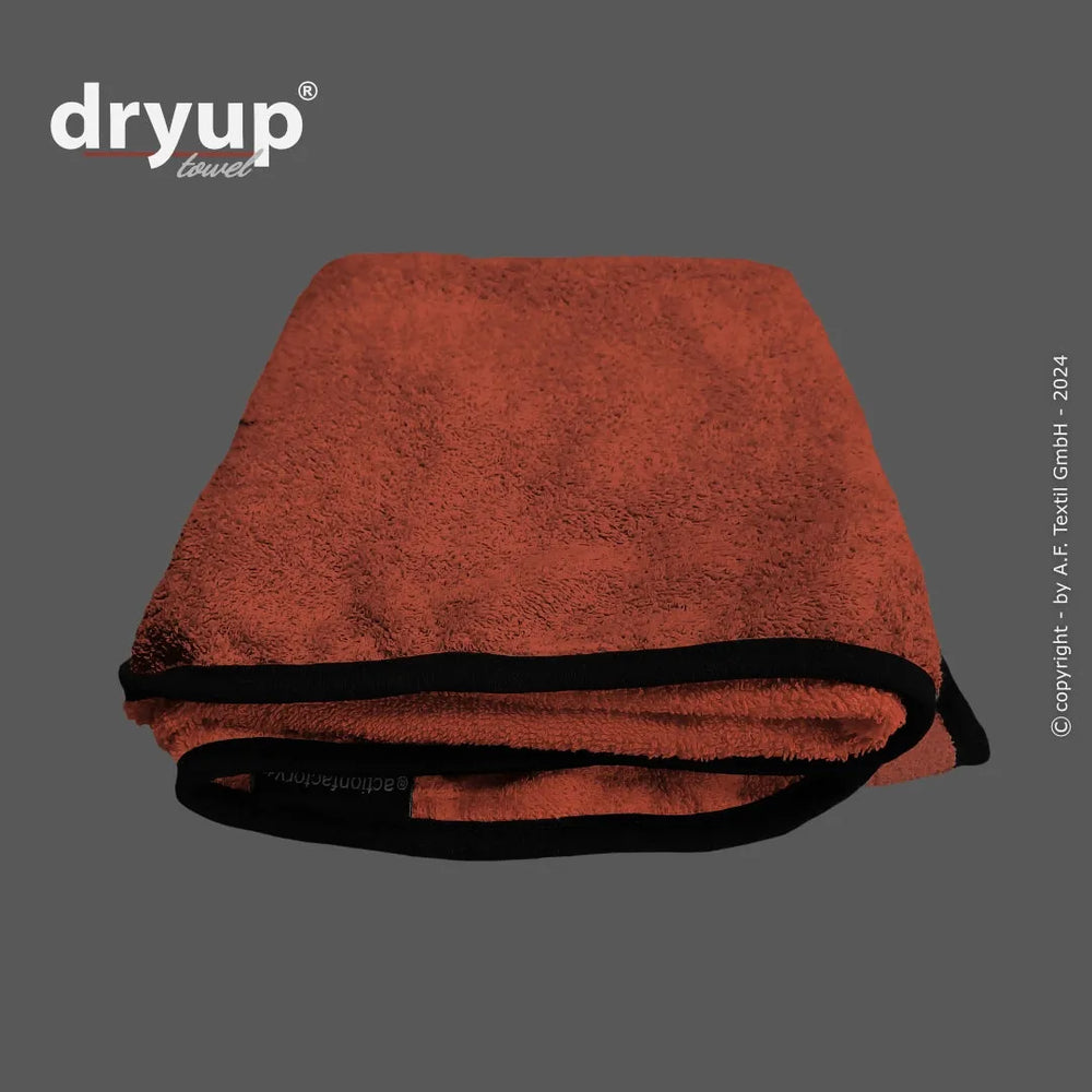 Zusammengeklapptes Dryup® Handtuch in warmem Orange mit schwarzem Saum auf grauem Hintergrund. Flauschiges Material, Markenlogo sichtbar.