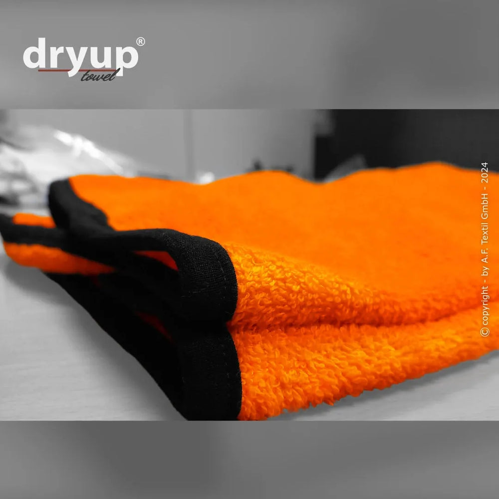 Orangefarbenes flauschiges Handtuch mit schwarzen Säumen, schräg auf einem Tisch platziert, mit „dryup“-Logo sichtbar.