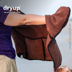 "Person hält braunes dryup® Handtuch mit Ärmel in natürlicher Umgebung am Strand, samtige Oberfläche und schwarzer Rand."