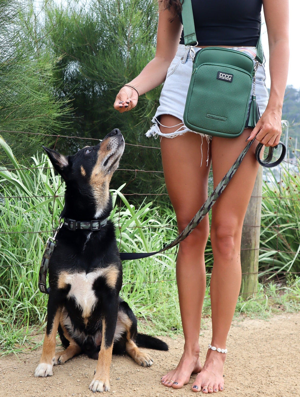 Frau mit Jeans und schwarzem Oberteil trägt grünen Umhängebeutel in der Natur und hält einen Snack in der Hand.