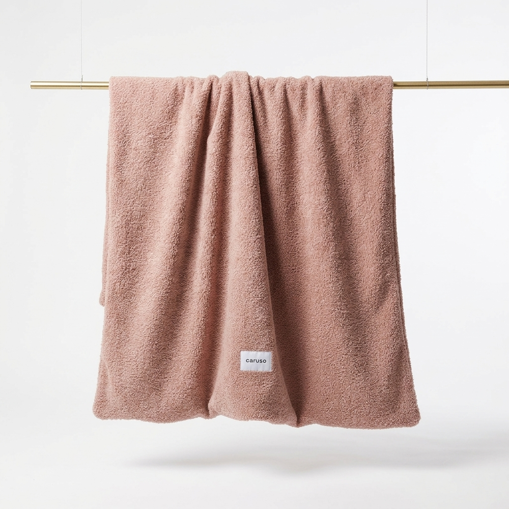 Weiches, rosafarbenes Teddy-Handtuch mit flauschiger Textur und weißen "caruso" Label, hängend an einer Stange.