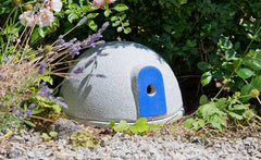 Graue kuppelförmige Struktur aus steinähnlichem Material im Garten mit blauer Öffnung und grüner Vegetation.