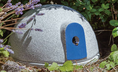 Graues Insektenhotel mit blauer Tür in grüner Umgebung, umgeben von Pflanzen und Lavendel. Ideal für nützliche Insekten.