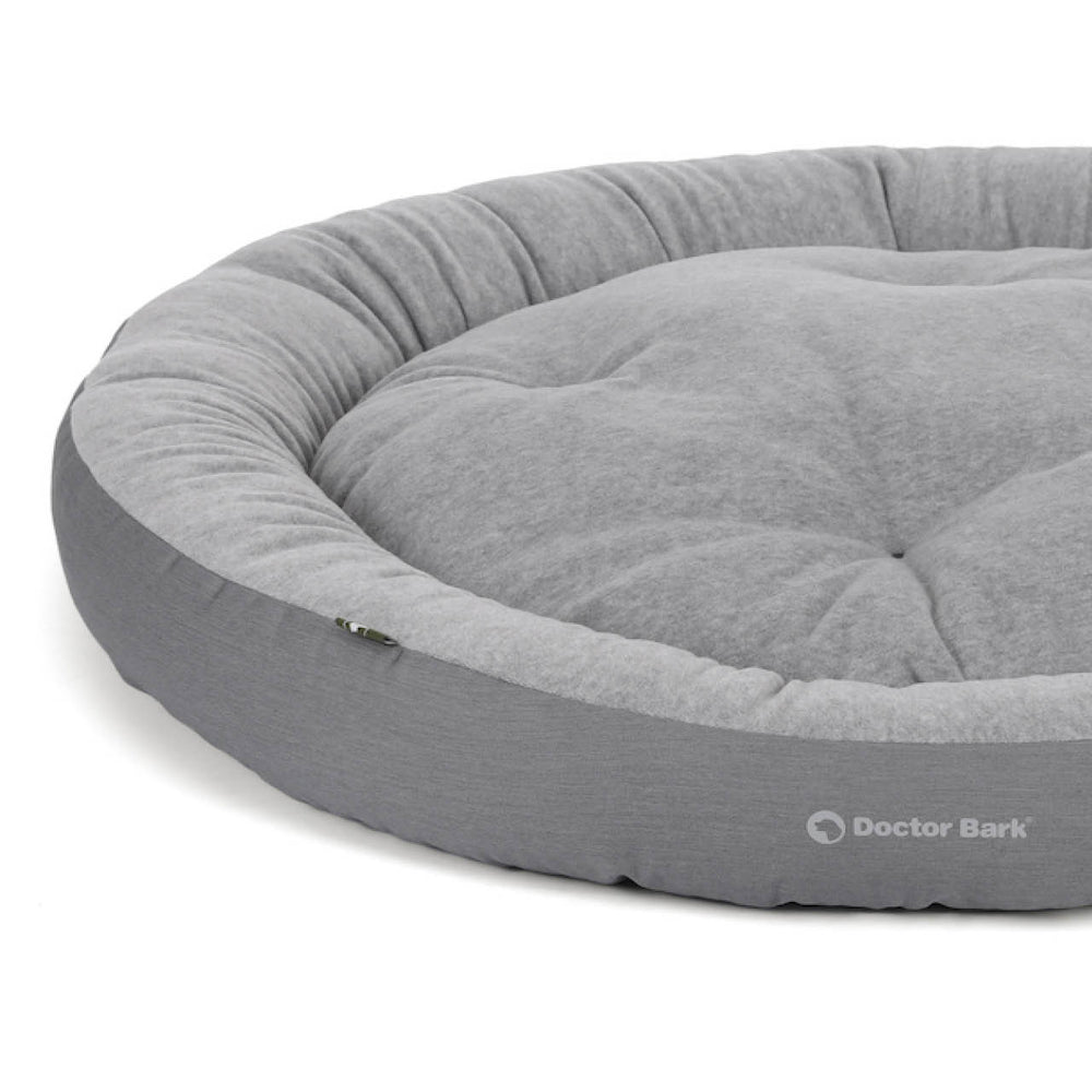 Gemütliches rundes Hundebett in hellem Grau mit gepolsterter Oberfläche und Doctor Bark Logo am Rand.