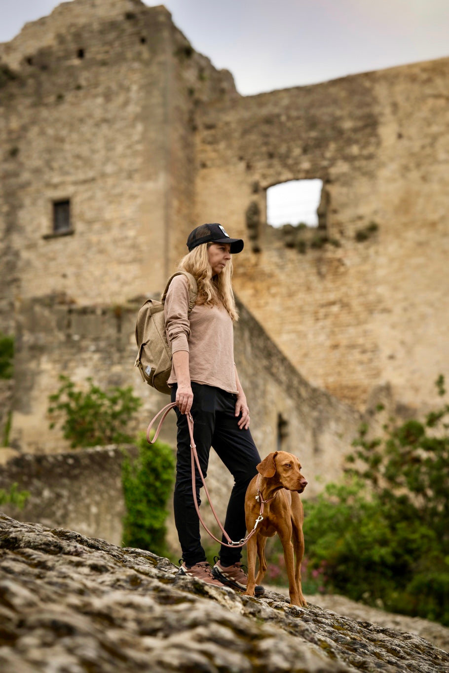 Frau mit Hund an Lederleine 2m, Hüte tragend, vor Burgruine und grüner Natur auf felsigem Gelände.