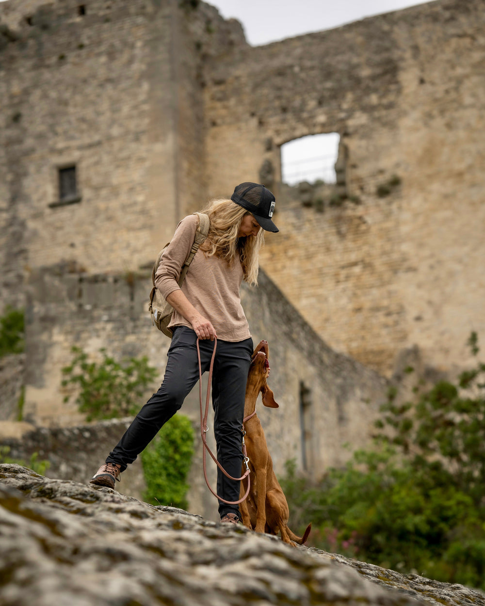 Person mit Hund an einer runden Lederleine in Gold an einem steinigen Hang vor einer alten Ruine.
