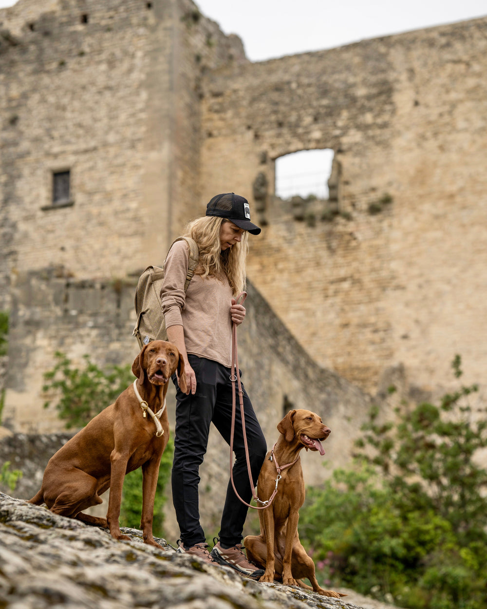 Frau mit blonden Haaren in Sportkleidung hält eine 2m lange Lederleine für Hunde vor einer Steinmauer.