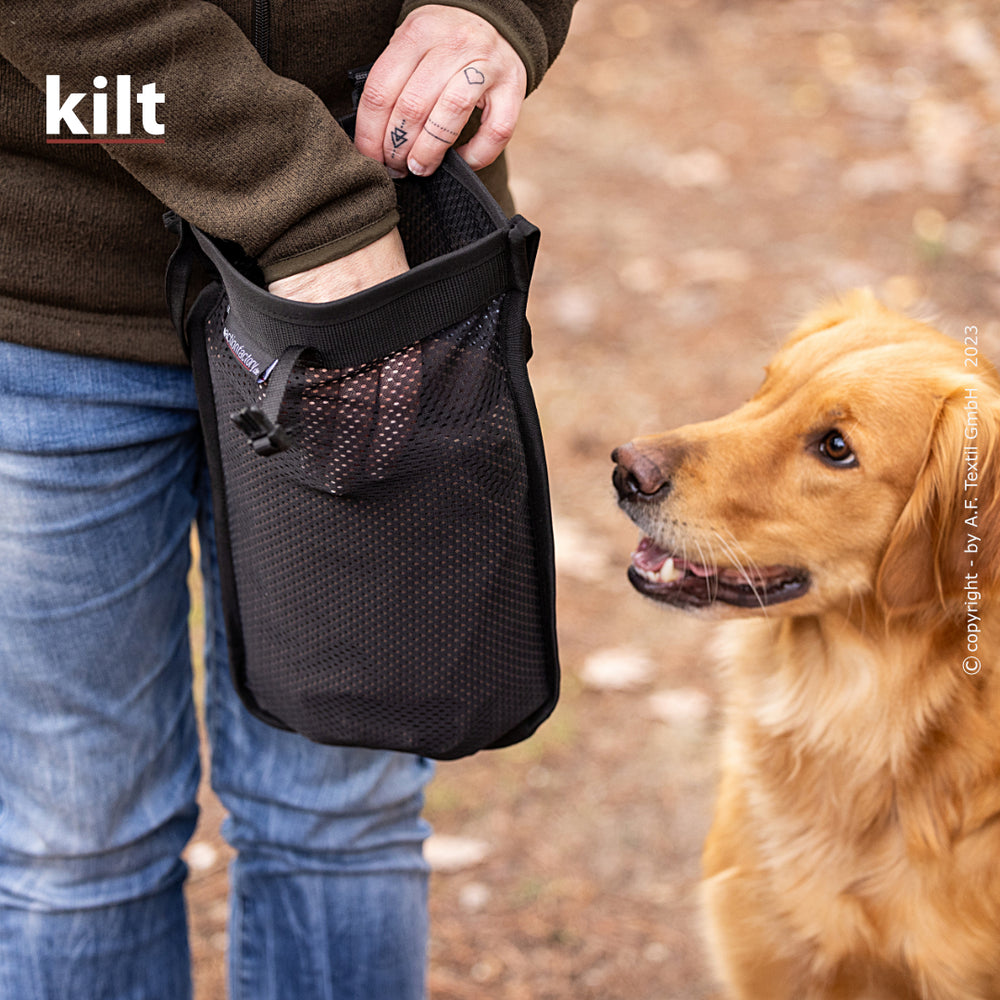 Person hält schwarzen Behälter mit Netzoberfläche, daneben goldenes Retrieverspielzeug auf dem Boden.