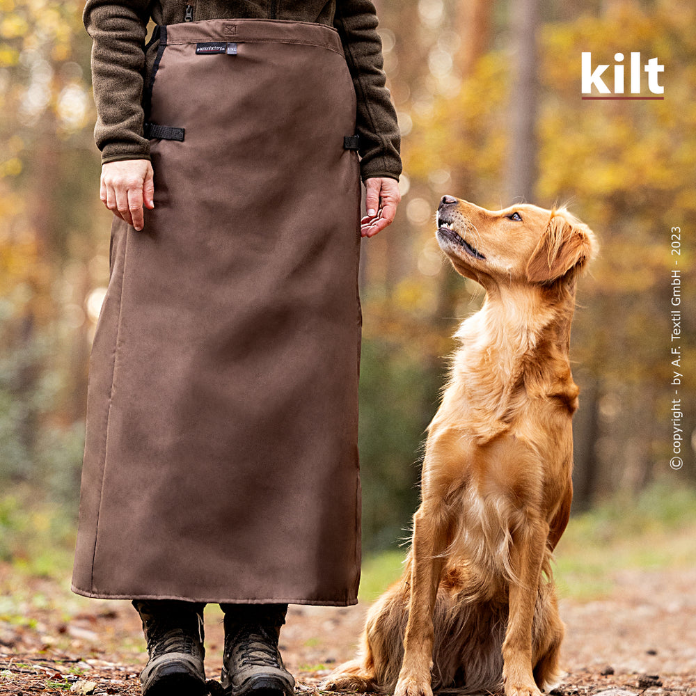 Person im braunen Kilt im Wald mit golden retrieverähnlichem Hund, umgeben von unscharfen herbstlichen Farben.