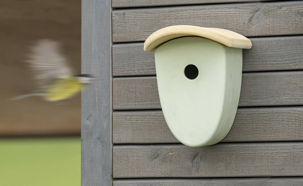 Nistkasten für Vögel an grauer Wand, mit runder Öffnung und abgerundetem Dach, während ein Vogel im Hintergrund fliegt.