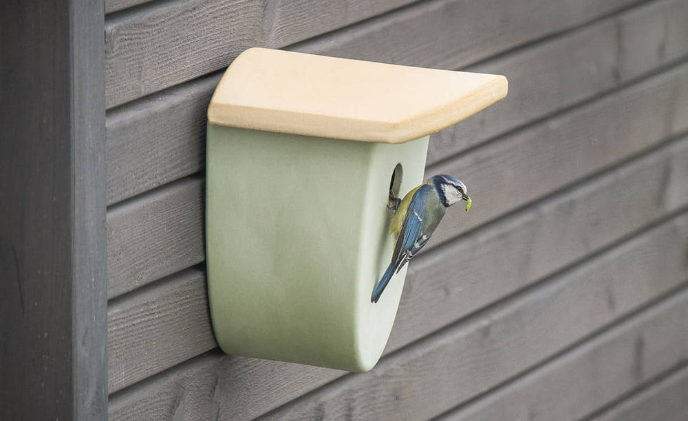 Modernes Vogelhaus in sanften Farben mit einer Meise am Eingang, vor einem dunklen Holz Hintergrund.