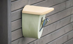 Modernes Vogelhaus in sanften Farben mit einer Meise am Eingang, vor einem dunklen Holz Hintergrund.