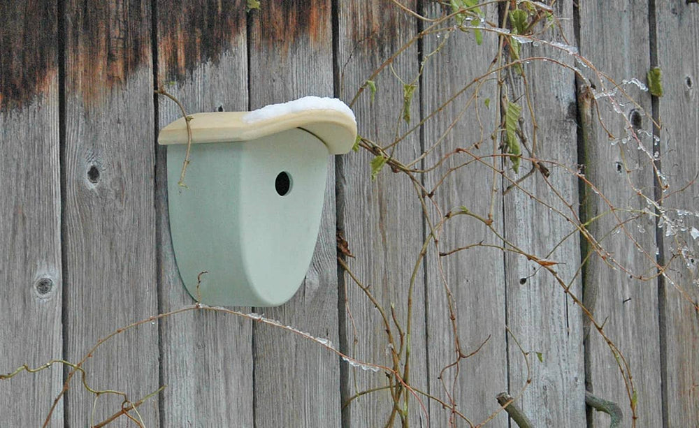 Vogelnest aus Kunststoff an einer Holzwand mit abgerundeter Form und heller Dachabdeckung, umgeben von Eiszapfen.