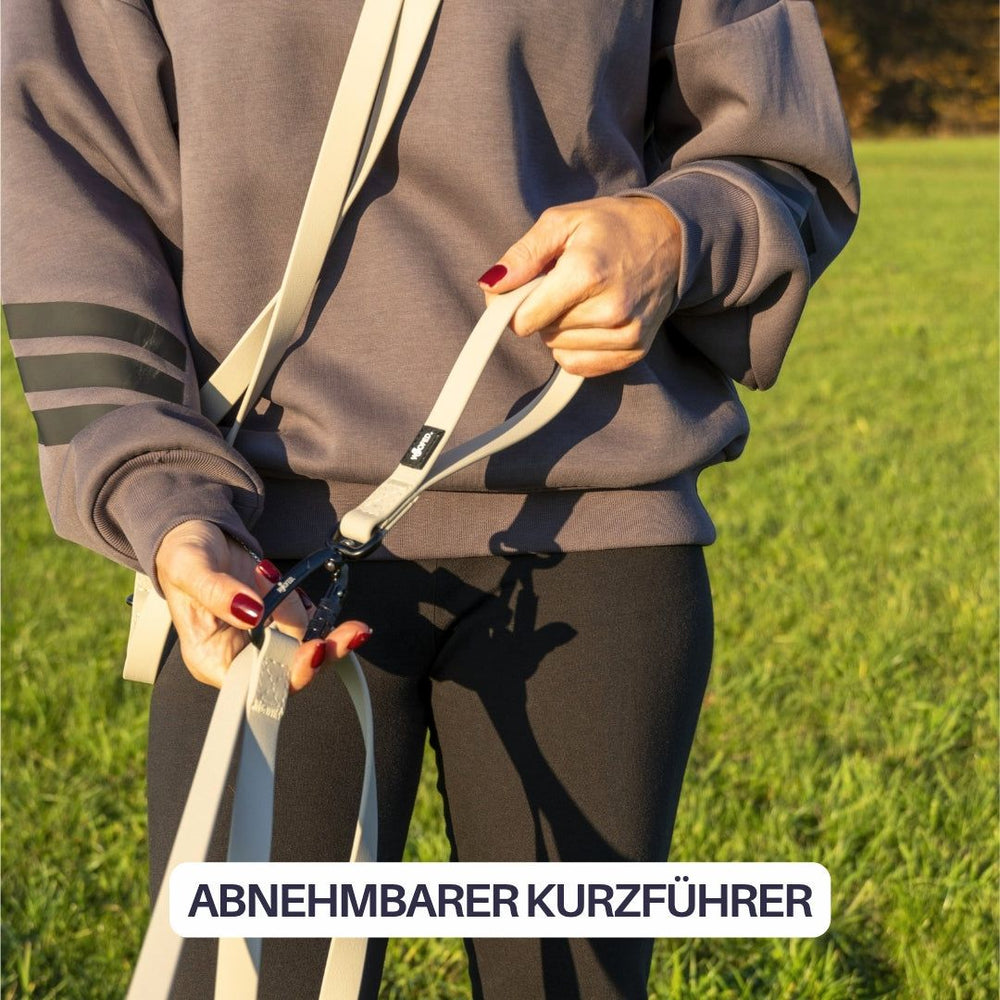 Person mit Kurzführer in grau gestreiftem Sweatshirt auf einer grünen Wiese, präsentiert die WOOFLEX® Schleppleine.
