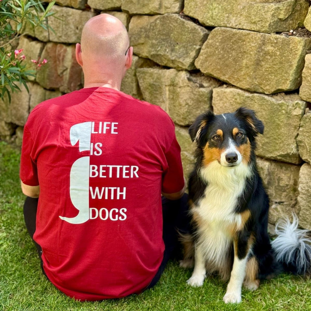 Mann mit Glatze sitzt in rotem T-Shirt "LIFE IS BETTER WITH DOGS" neben einem schwarzen Hund.