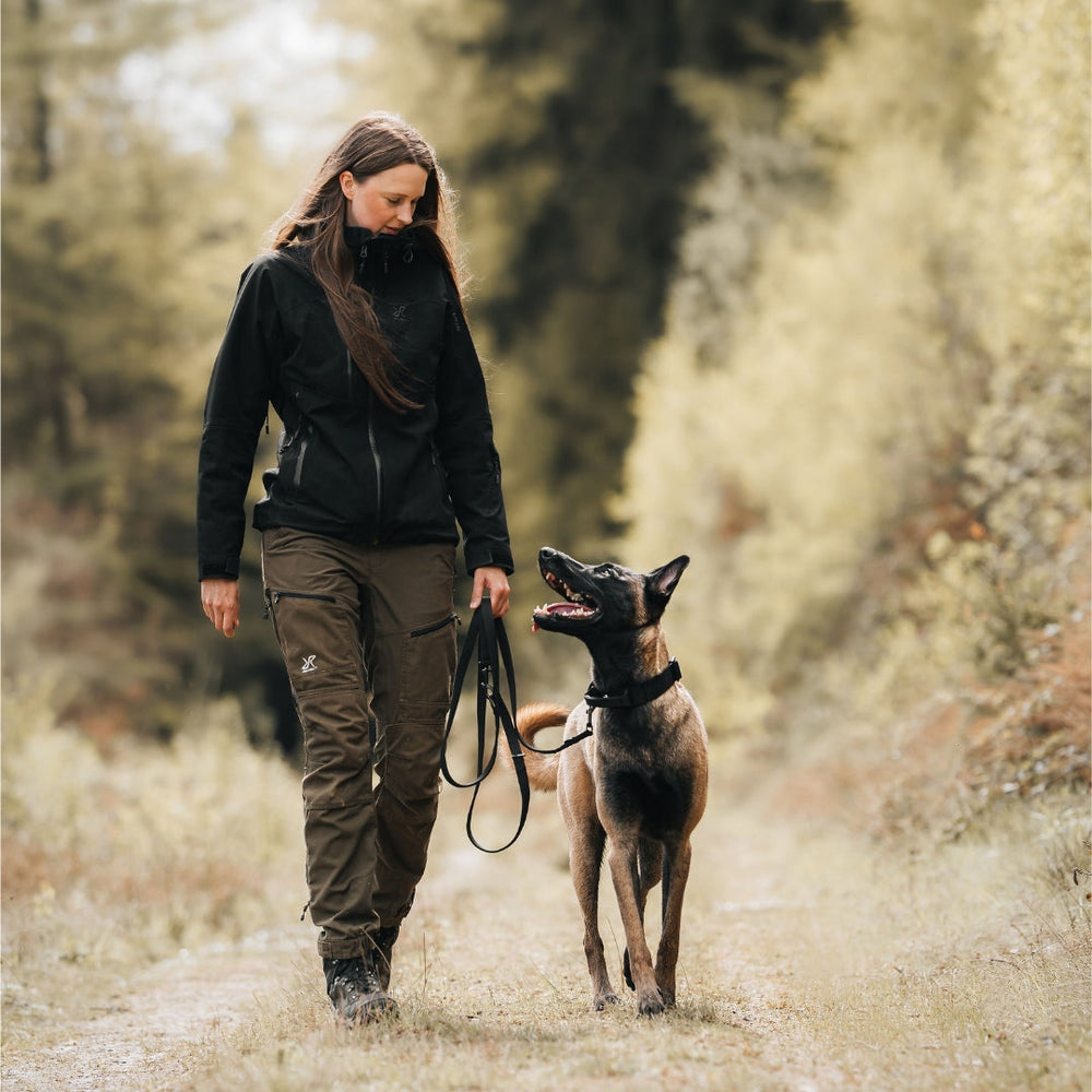 Frau mit schwarzer Jacke und olivgrünen Hosen spaziert mit belgischem Malinois auf einem Waldweg.
