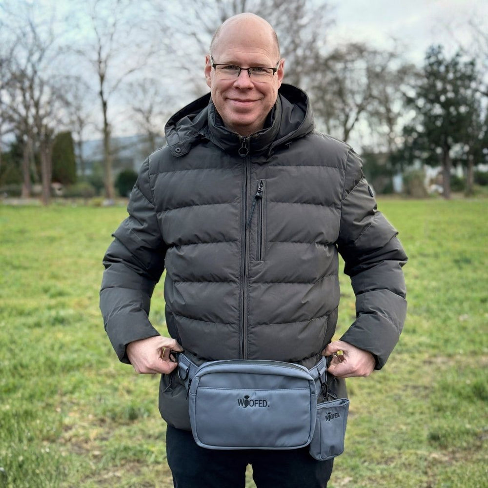 Mann im grauen Daunenmantel trägt graue multifunktionale Gassitasche als Bauchtasche in einer grünen Umgebung.