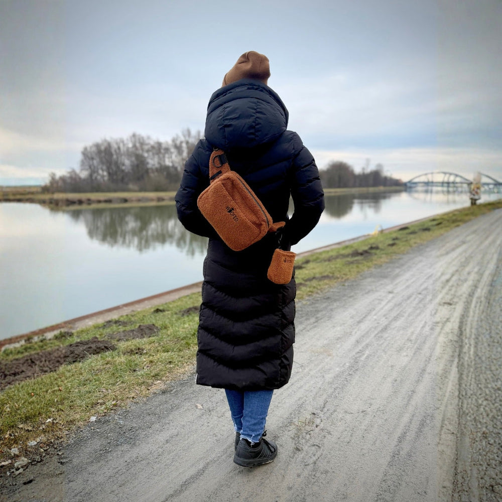 Person am Gewässer mit langer, schwarzer Daunenjacke und brauner Mütze, trägt multifunktionale Gassitasche.