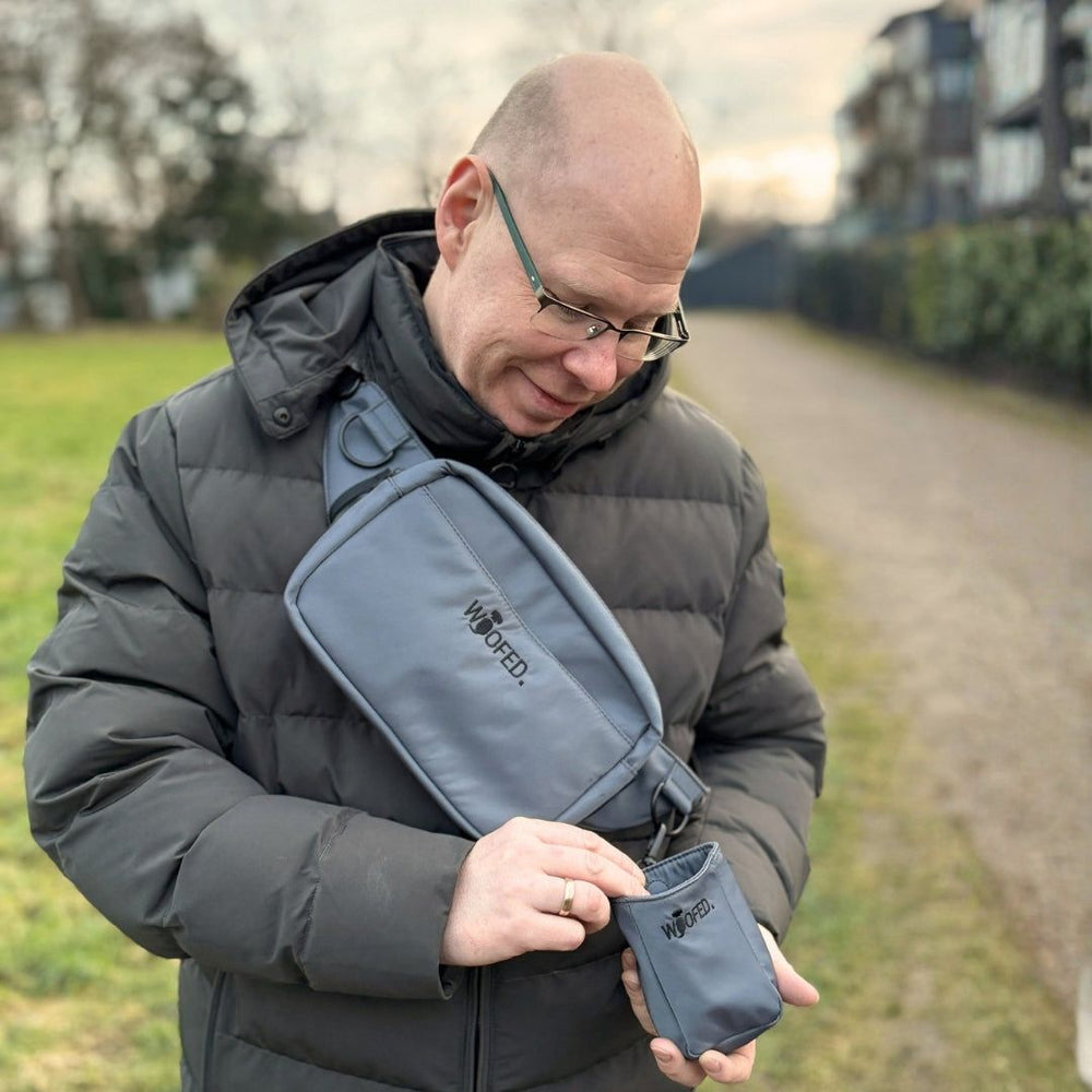 Mann mit grauem Parka hält graue WOFED Umhängetasche und schaut in die kleine Tasche auf einem Weg im Freien.