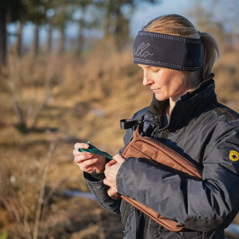 Frau im Wald mit dunkelblauem Kopfband und Jacke, hält braune multifunktionale Gassitasche.