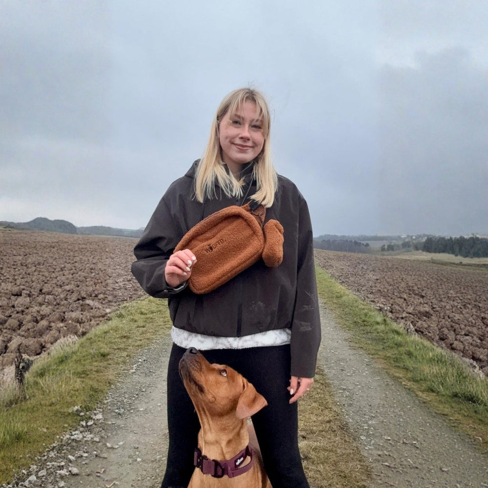 Frau mit brauner Gassitasche auf Feldweg, Hund sitzt neben ihr, bewölkter Himmel im Hintergrund.