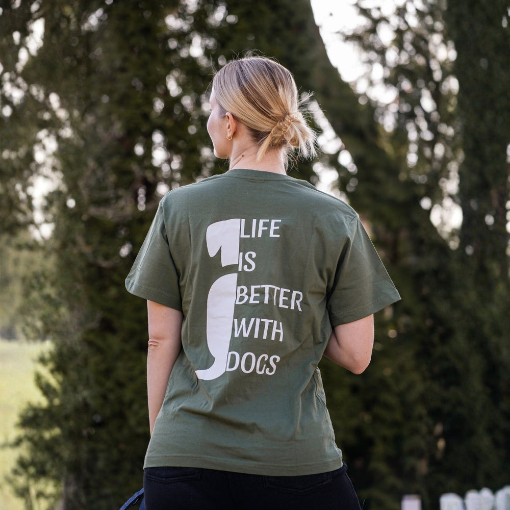 Person in grünem T-Shirt mit Aufdruck "Life is better with dogs" steht vor Bäumen in der Natur.