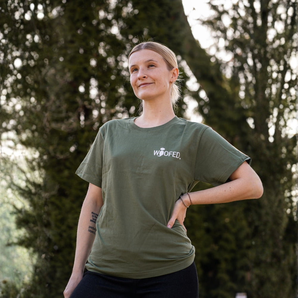 „Person mit olivegrünem T-Shirt 'WOOFED' in natürlicher Umgebung mit Bäumen, stolz und selbstbewusst stehend.“