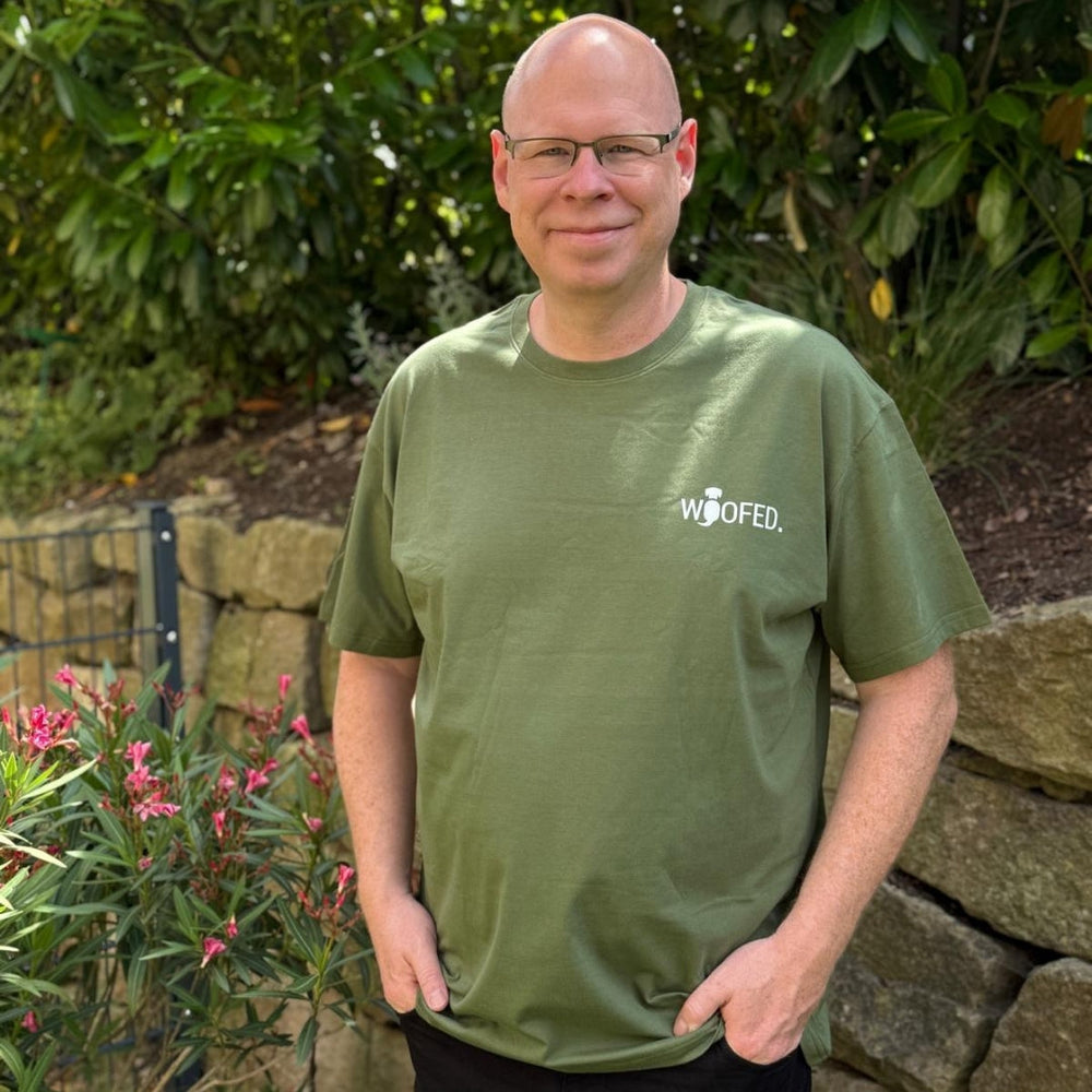 Mann mit Glatze lächelt im Garten, trägt olivgrünes T-Shirt "Life is better with dogs".
