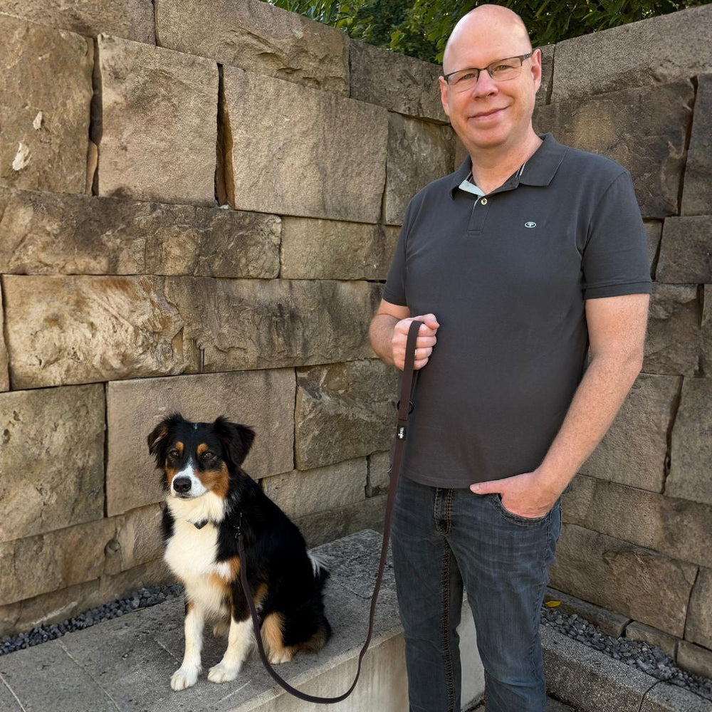 Mann mit Brille hält WOOFLEX® Pro Führleine neben schwarzem und braunem Hund vor steiniger Wand.