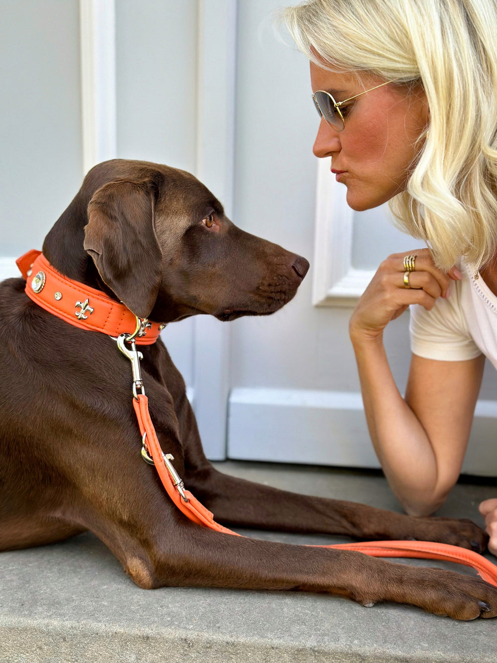 Frau mit lockigen blonden Haaren beugt sich zu ihrem braunen Hund mit orangefarbenem Halsband in der Sommer Edition.