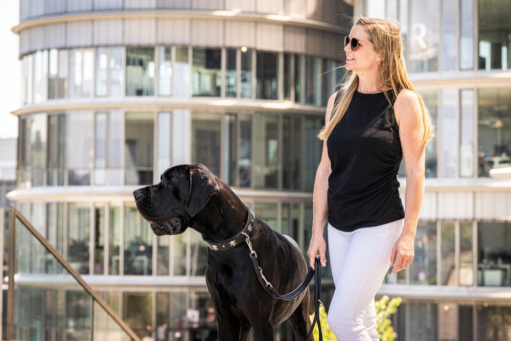 Frau in schwarzer Weste mit Hund an der Leine, trägt Sonnenbrille, moderner Hintergrund.