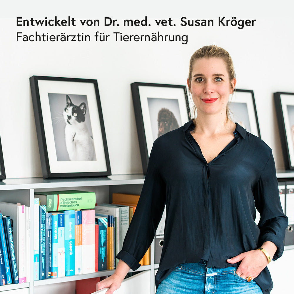 Frau mit blonden Haaren in modernem Raum, schwarzes Hemd, umgeben von Tierbildern und Bücherregalen.