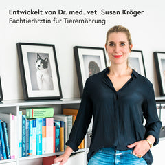 Frau mit blonden Haaren in modernem Raum, schwarzes Hemd, umgeben von Tierbildern und Bücherregalen.