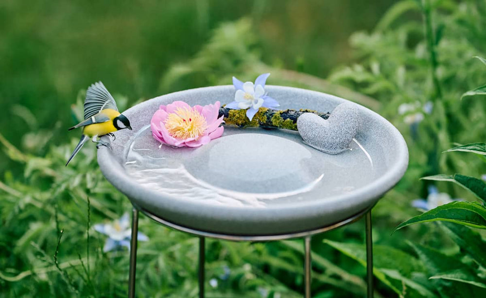 Vogeltränke Granicium auf Ständer, gefüllt mit Wasser, dekoriert mit Blume und Herz, kleiner Vogel am Rand.