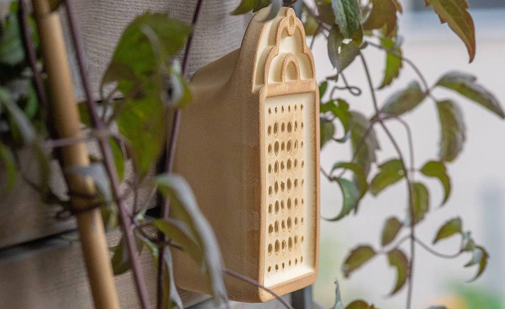 Beige Wildbienenhaus an Wand, umgeben von Pflanzen, mit strukturierten Löchern für Insekten.