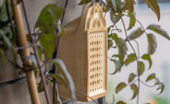 Beige Wildbienenhaus an Wand, umgeben von Pflanzen, mit strukturierten Löchern für Insekten.